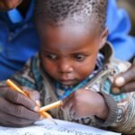 A young child carefully holds a pencil while learning to write under the guidance of an adult.